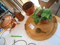 Copper items including kettle, two planters (one with artificial plant), copper pot with stand on white tiled surface, wood tray partially visible.