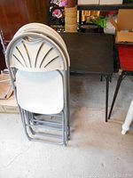 Photo showing black folding card table with metal frame and four stacked folding chairs with white plastic seats and backs.