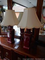 Pair of red ceramic table lamps with beige fabric shades, placed on a wooden surface in front of large mirrors.