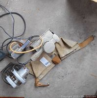 Three corded power tools with taped cords and visible wear, sitting on concrete floor alongside a leather tool pouch.