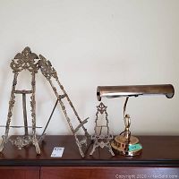 Two large standing brass frames next to smaller brass standing frame and brass desk/piano lamp shown on wooden surface against neutral wall.