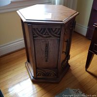 Photo of hexagonal solid wood end table showing decorative carved wood pattern on door panels and brass-tone handle on hardwood flooring.