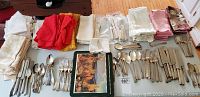 Top-down view of all flatware and napkins spread out showing quantity and variety of silver plate forks, knives, spoons, and multiple color cloth napkins.