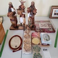 Photo of three papier mache figurines, sets of floral-shaped candles, a small oval frame, and a brooch box on a table.