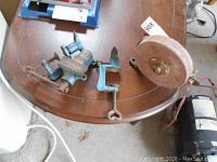 Blue 2.5 inch hobby vise and two blue clamp-on hobby anvils on a wooden table with an older hand crank sharpening stone visible.