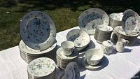 Full set arranged on a white tablecloth outdoors showing stacks of plates, cups, bowls, and serving pieces all with a floral pattern accented with gold rims.
