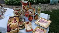 Overview of the complete Tuscan style poppy hostess set, showcasing the ceramic pitcher, jar, oil drizzlers, glass cups, plates, and themed towels on a white tablecloth outdoors.