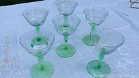 Six green stemmed cordial glasses arranged in two rows on a white cloth. One glass shows a crack on the bowl.