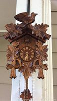 Front view of wooden cuckoo clock showing carved bird on top, leaf decorations, Roman numeral clock face with hands, and pinecone weights hanging below.