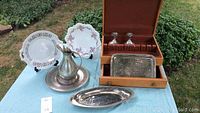 Wide shot of all items on display on a blue tablecloth outdoors, showing the pewter pitcher and tray, hammered tray, wooden silverware box with silverware and two glass decanters, and two decorative plates.