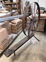 Photo of a large, antique wooden spinning wheel with a vertical wheel and wooden base, surrounded by cardboard boxes in a storage setting.