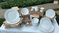 Various cream-colored china pieces arranged in boxes and stacks on ground, showing plates, bowls, cups and napkin rings.