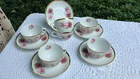 Five tea cups and matching saucers arranged on a white crocheted tablecloth outdoors, showing pink rose floral pattern with gold rims.