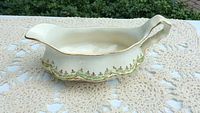 Full side profile of cream-colored ceramic gravy boat with gold trim and lime green floral detailing on base edge, placed on a crocheted lace cloth outside.