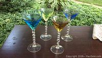 Four different colored hand blown stemware glasses with twisted clear stems on a dark surface, outdoors with greenery background.
