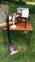 Overview of items on triangular folding table outdoors on grass including Dirt Devil vacuum, paper towel holder, doll, iron, fabric bin, wooden stool, basket, and spring form rod box.