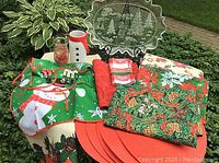 Photo showing the Christmas themed tablecloths, placemats, napkins, snowman jar, and child apron all arranged together on grass outdoors.
