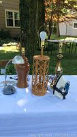 Photo showing three lamps including wooden base lamp, lamp with rattan base, brass-tone lamp base, also showing metal dome-shaped shade and Proctor Silex iron on a white table outdoor.