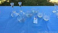 Overview of all items including two taper candle holders, hexagon glass bowls, shot glass and patterned highball/old fashioned glasses on a blue tablecloth, outside.