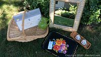Full view of lot items arranged on grass: basket holding sheet and green ceramic pitcher, mirror leaning on basket, tole tray with floral painting, wooden tray with two tea cups