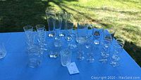 Group of multiple clear bar glasses placed on blue cloth with grass in background. Includes various types and sizes, some with branding.