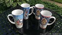 Four winter-themed extra tall ceramic hot cocoa cups displayed on a black table outside. Each cup features colorful winter scene artwork including houses and snowmen.