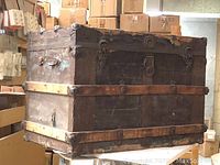 Front and top side view of vintage wooden trunk showing metal trim, wood slats, metal latches and leather handle.