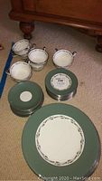 Stack of 15 dessert and dinner Wedgwood plates with green border and a set of 12 cups and saucers arranged on carpet.