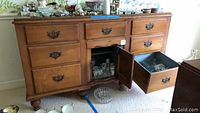 Wood buffet with multiple drawers, central cabinet door open showing glassware inside. Various items on top of buffet. Scratches visible on wood surface.