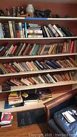 Wide shot of multiple bookshelves filled with many books in hardcover and paperback formats, mostly upright and some stacked horizontally. Small figurines and decorative items on top shelf.
