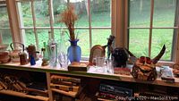 Wide shot displaying various glass vases, ceramic teapot, brass pitcher, papers, mini building, and decorative mask on window ledge in natural light.