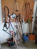 Wide view of assorted yard tools hanging on pegboard including rakes, pitchfork, shovel, saws, and mop