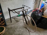 Metal garden cart placed on concrete floor in garage with large single spoked wheel and open rectangular frame. Visible rust on metal surfaces.