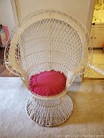 Front view of white wicker peacock chair with red fringed cushion placed on beige carpet.