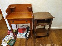 Two wooden furniture pieces side-by-side on a wooden floor next to wallpapered wall: a small desk with a single drawer and back ledges, and a darker side table with two shelves and turned legs. Scratches visible on surfaces.