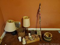 Two table lamps with beige shades, woven baskets, cups, a wooden walking stick decorated with ribbons, a rectangular ornate tissue box holder and a small metal alarm clock.
