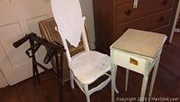 Photo showing the white painted chair with ornate high back, wooden hamper with leather straps, and side table grouped together.