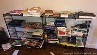 Wide view of the black metal book stand filled with various books on three shelves against a wall on a wooden floor.
