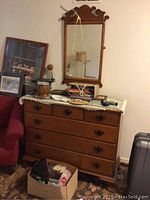 Wooden dresser with four drawers, metal handles, and decorative items on top, next to a red armchair and some framed pictures.