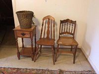 Full lot showing two wooden chairs, side table with drawer and lower shelf, and woven wicker basket on top of table.
