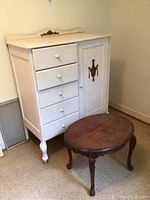 Wide shot showing the white cabinet with five drawers, a door with brass hardware, and a decorative gold ornament on top. In front is the dark wood oval coffee table.