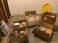 Four cardboard boxes with packed dinnerware and glassware, some wrapped in newspaper, in a basement room