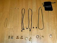 All jewelry items including silver necklaces, black bead necklaces with earrings, silver bracelet, cufflinks, and pendants arranged on a wooden surface.