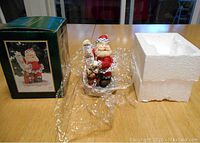 Santa Claus Christmas figurine beside its original box and styrofoam insert on a wooden table.