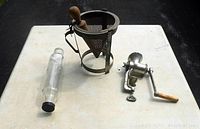 Glass rolling pin, metal cone strainer with wooden pestle, and vintage metal meat grinder laid out on a white surface.