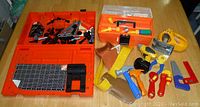 Image shows the full set of toys including the orange toolbox and assorted toy hand tools like hammers, saws, clamps, and drill in varied colors scattered on a surface.