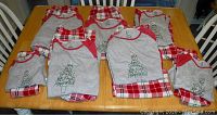 7 pairs of folded Christmas pajamas arranged on wooden table showing grey tops with text Christmas tree design and red plaid pants