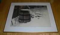 Front view of the black and white framed photograph showing a wooden barrel in the snow next to a wooden wall.