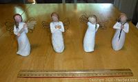 Four Willow Tree figurines in a row on a wooden table with measuring ruler for scale.