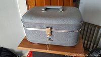 Gray vintage McBrine vanity case closed, sitting on a wooden table, visible top handle and latch lock with key attached.